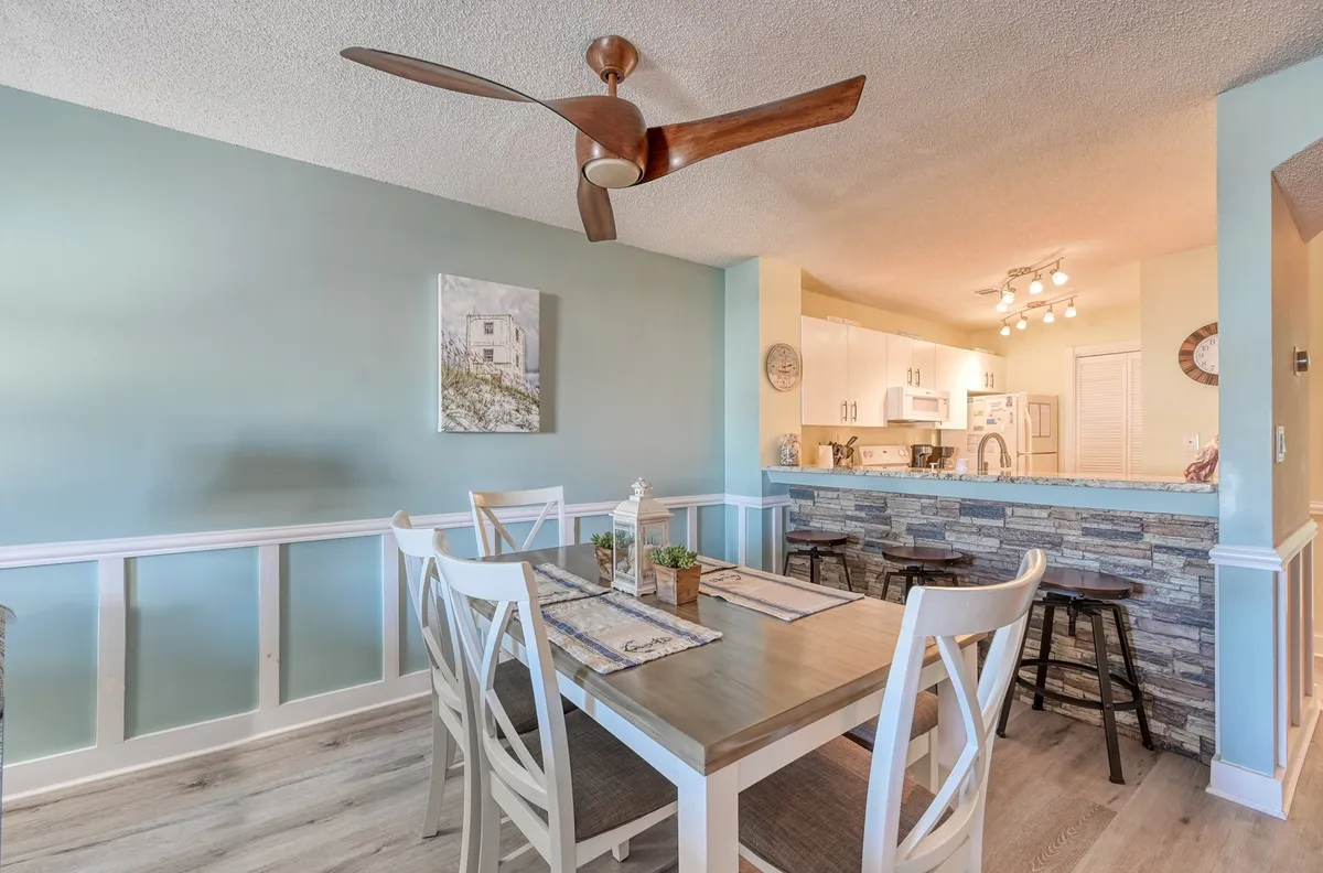 A dining area with a table set for six, ceiling fan above, artwork on the wall, and adjacent kitchen bar area. The space is inviting and designed for shared meals, game nights, or planning future beach adventures, mirroring the section's focus on gathering.