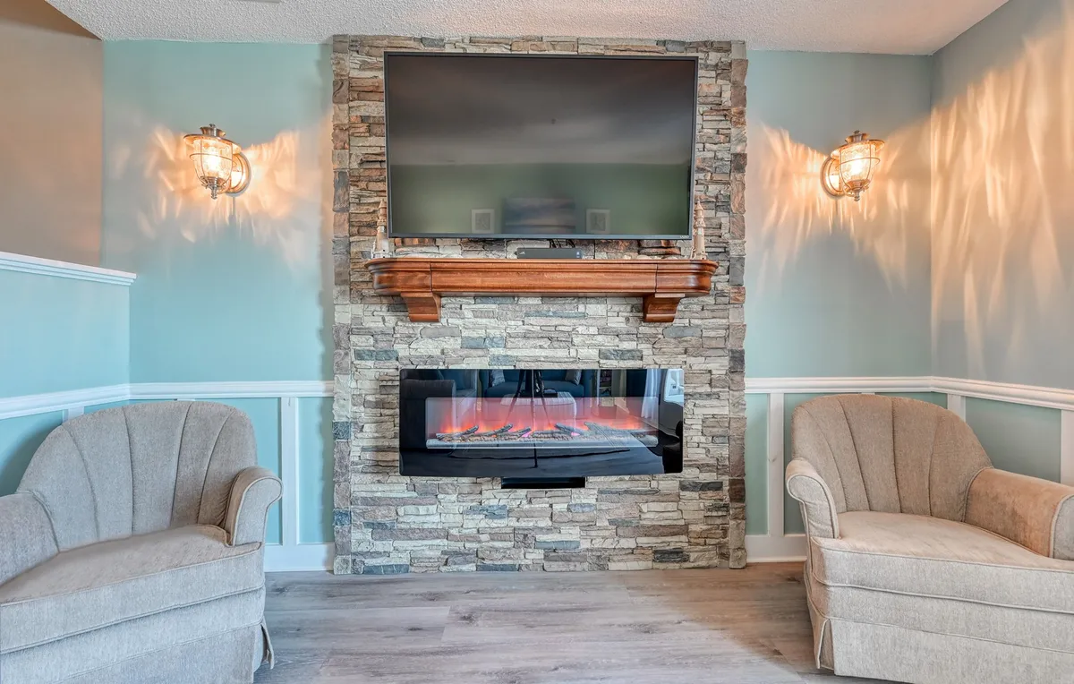 A warm, modern living area featuring a stone fireplace, modern television, and comfortable armchairs. The ambiance is inviting, ideal for relaxing together after a day by the pool or at the beach.