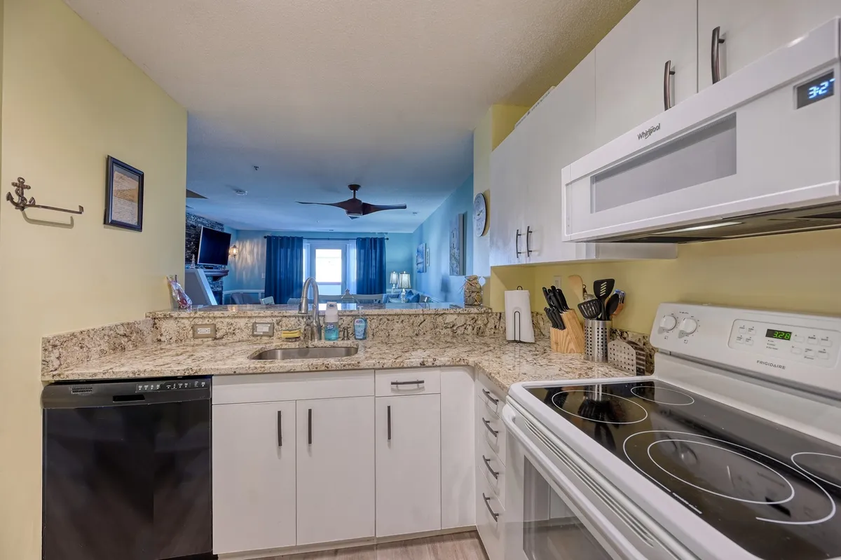 This image features a welcoming kitchen space with a granite countertop, white appliances, and a spacious sink. The scene captures ongoing activities such as cooking and entertaining, and hints at family gatherings around the kitchen island, with an overall mood that is bright and inviting.