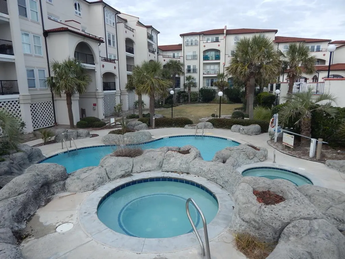 The image showcases two inviting jacuzzi pools set among rock formations and palm trees, with the resort's buildings in the background. The area is designed for year-round relaxation and socializing.