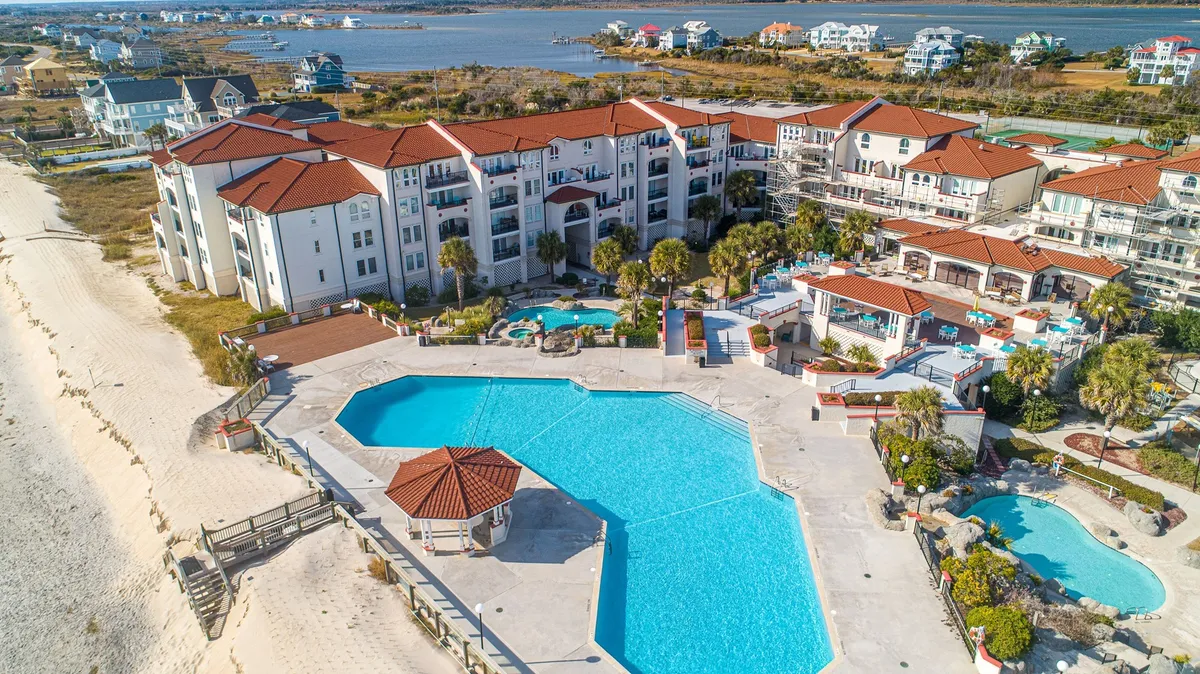 A wide exterior view capturing the resort's multiple swimming pools surrounded by landscaped gardens and framed by beachfront buildings. The pools are the focal point, with inviting turquoise water and sun loungers, hinting at both relaxation and family fun. The ocean is visible just beyond, seamlessly connecting the pool area to the beach.
