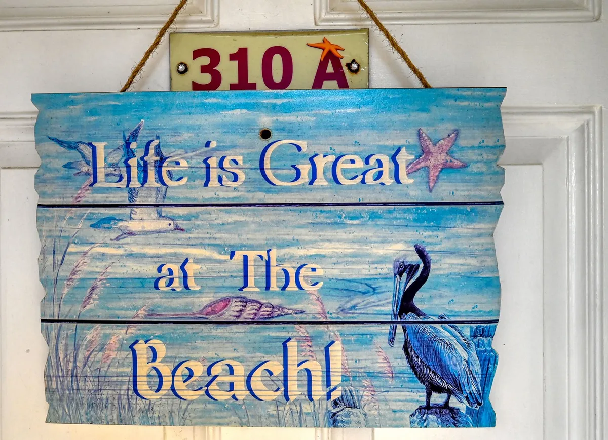 A cheerful, sunlit interior space featuring a decorative sign with text, an illustration of a pelican, and scattered starfish and shells. The ambiance hints at the beach just beyond, evoking the feeling of seaside adventures and the local wildlife. The decor includes references to sunset views, beachcombing, and local seafood dining, subtly connecting the interior with the natural experiences outside.