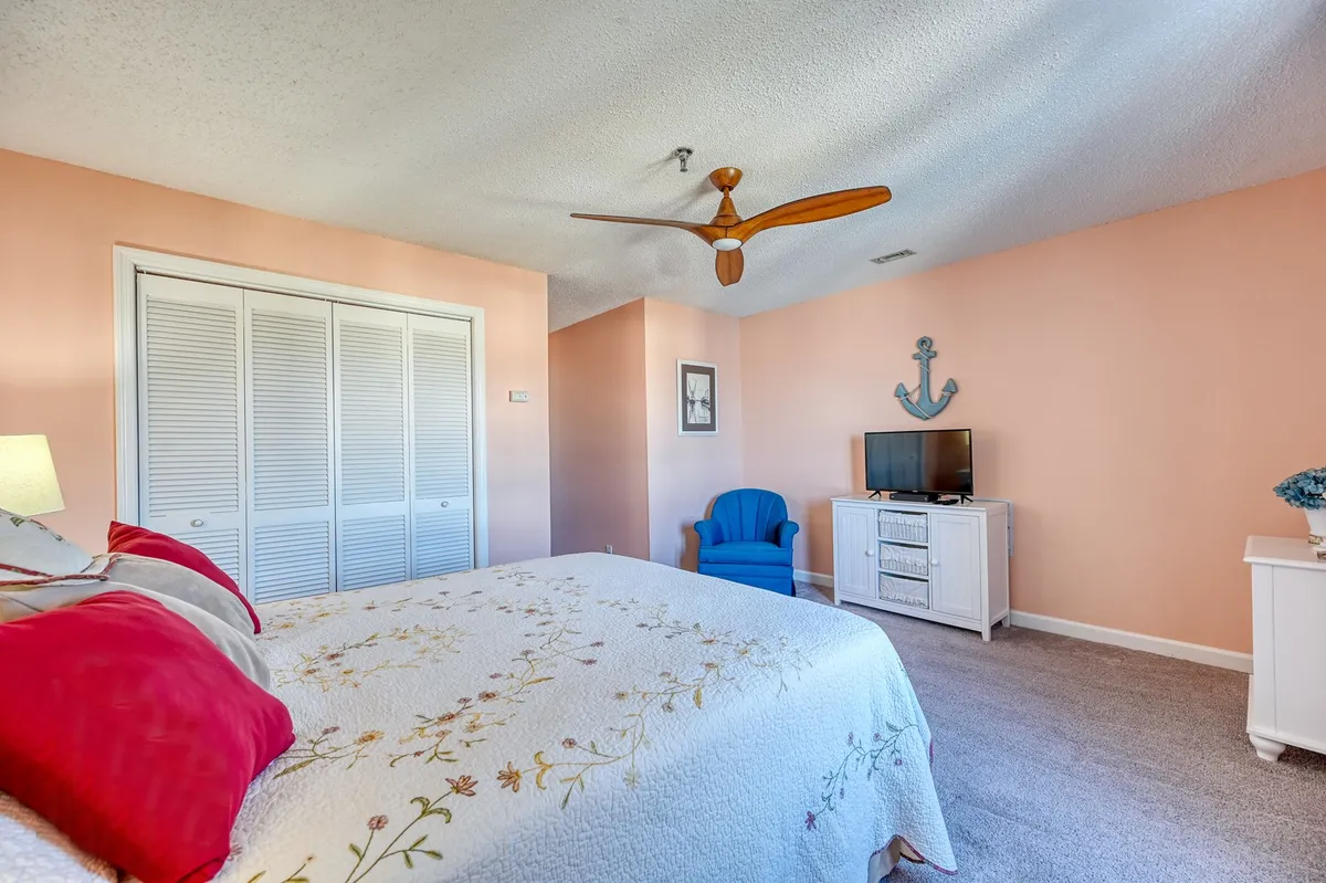 A comfortable bedroom with a queen bed topped with a floral quilt, a blue accent chair, and a TV unit with decor. Soft natural light filters in, creating a restful vibe perfect for unwinding after a day in the sun.