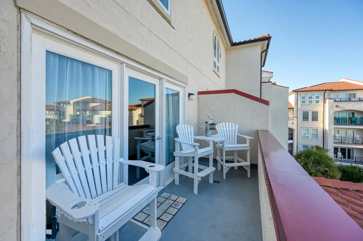 An inviting oceanfront balcony features white Adirondack chairs facing out toward the sunrise, with sliding glass doors leading back inside and a sturdy balcony railing. The tranquil scene suggests a peaceful start to the day, perfect for savoring coffee while taking in breathtaking beach views.
