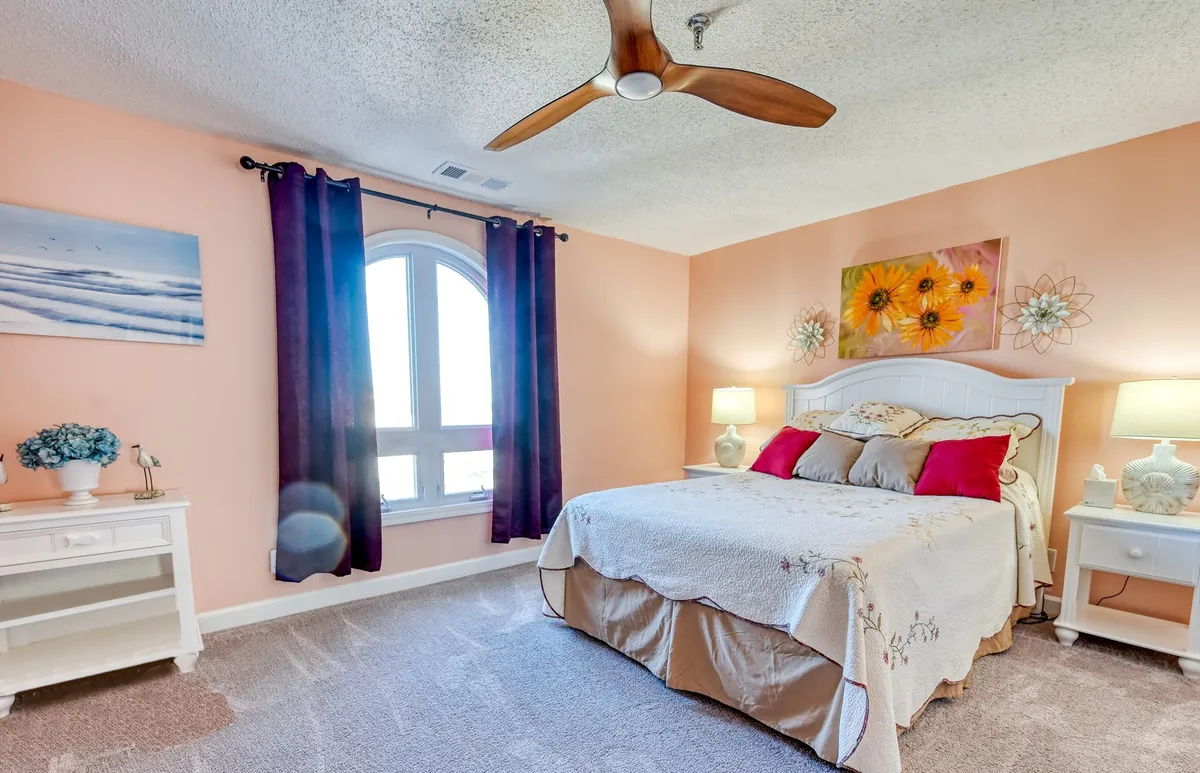 This image features a cozy bedroom with a queen bed adorned with decorative pillows, a striking arched window that allows abundant natural light, colorful artwork, and nightstands with lamps. The room exudes a welcoming, restful atmosphere ideal for relaxation.