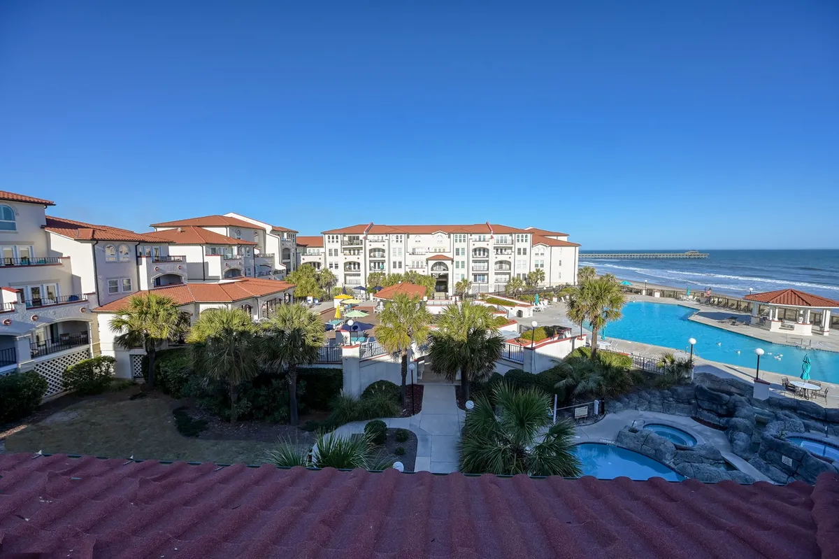 An inviting exterior shot of a swimming pool framed by palm trees and overlooking the ocean, with residential resort buildings in the background. The setting feels welcoming for both sunbathing and group family activities, with plenty of space for kids to play safely.