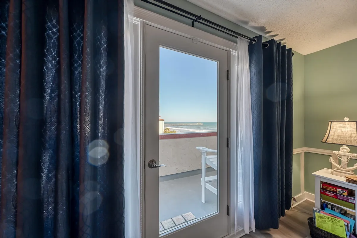 A serene interior view with a glass door leading to a balcony, ocean view visible, decorative lamp, and curtains softly framing the scene. The tranquil atmosphere is perfect for enjoying morning coffee or sunset cocktails while sea breezes drift in.