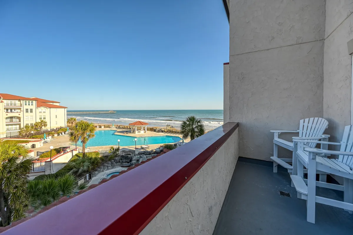 The hero image showcases a private balcony with classic Adirondack chairs overlooking a sparkling resort pool, swaying palm trees, and sweeping ocean views that stretch to a picturesque pier in the distance—an inviting scene for relaxation and coastal enjoyment.