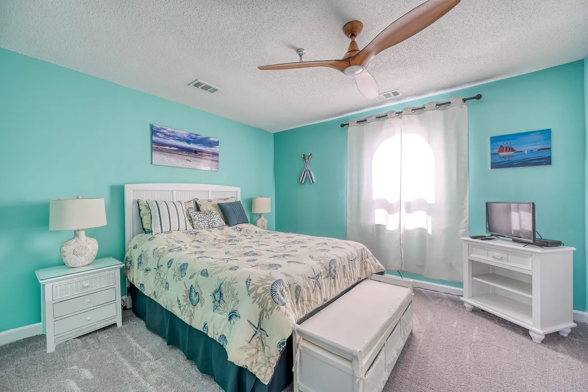 A peaceful bedroom featuring a neatly made bed with decorative coastal-themed bedding, white furniture, a ceiling fan, and calming wall art. The room is bathed in soft natural light, enhancing its restful ambiance.
