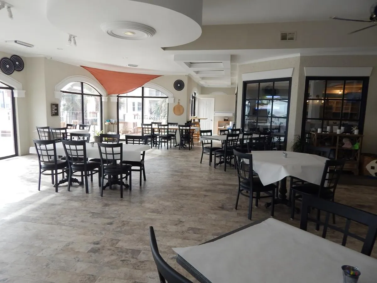 An airy, light-filled interior of the on-site restaurant, featuring tables with white tablecloths, black chairs, and an orange sail canopy above. Large windows frame relaxing views, creating an inviting atmosphere for dining and socializing.