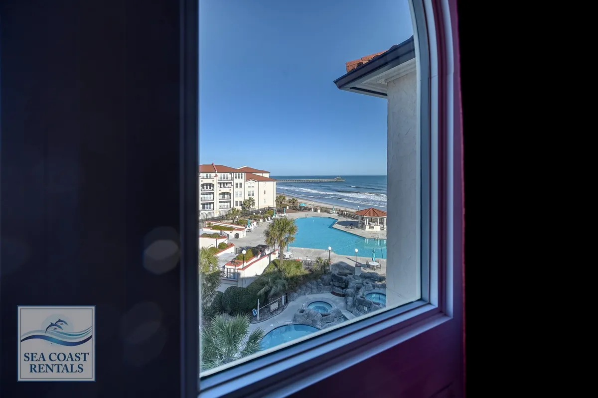 A view featuring a sparkling swimming pool with palm trees and the ocean in the background. The scene captures a lively, sunlit pool area ideal for swimming and socializing, with adjacent barbecue and seating spaces visible, suggesting versatility for family gatherings.